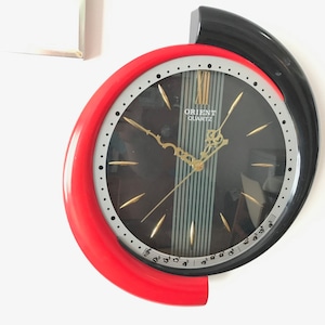 May include: A red and black wall clock with a silver face and black hands. The clock face has the words "Orient Quartz" printed on it.