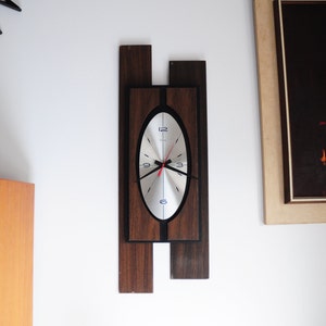 May include: A mid-century modern wall clock with a silver face and black hands. The clock is framed in dark wood with a rectangular shape.