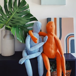 May include: Two plush dolls, one light blue and one orange, are embracing on a black shelf. The blue doll has orange arms, and the orange doll has blue arms. A large green monstera leaf and a white ceramic jug are to the left.