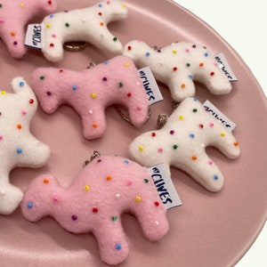 May include: Keychains in the shape of a camel, in pink and white felt, are scattered on a pink plate. The camels are decorated with colorful, embroidered dots. Each keychain has a small tag that reads "MY CLIVES".