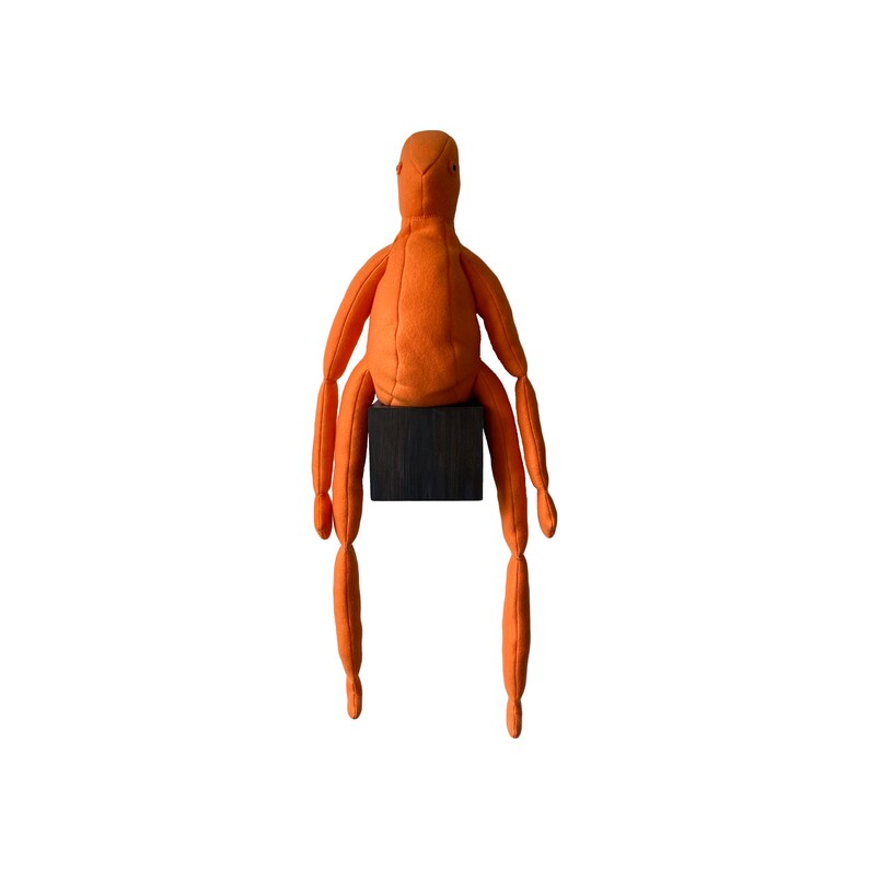 May include: An orange fabric doll with articulated limbs and a rounded head, seated on a small, dark wooden block. The doll's arms and legs are long and slender, giving it a unique, stylized appearance. The background is plain white.