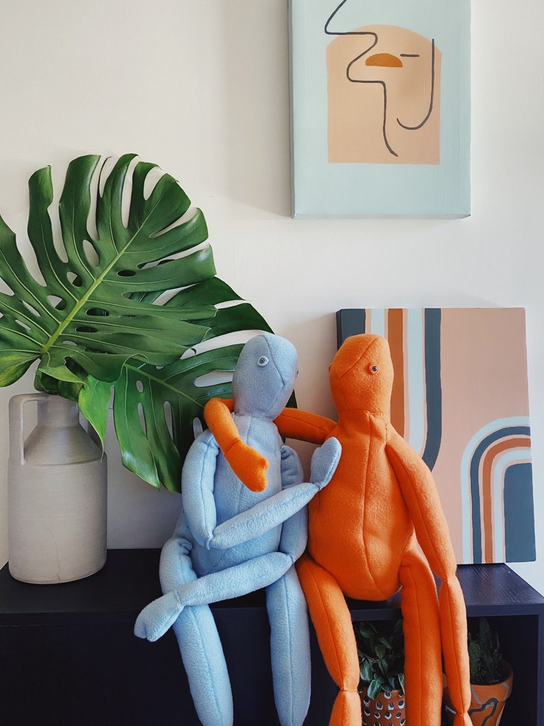 May include: Two plush dolls, one light blue and one orange, are embracing on a black shelf. The blue doll has orange arms, and the orange doll has blue arms. A large green monstera leaf and a white ceramic jug are to the left.