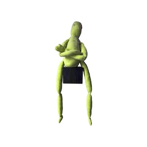 May include: A lime green fabric doll with a seated pose. The doll's arms are crossed, and it sits atop a small, dark wooden block. The doll's legs dangle, and the background is white.