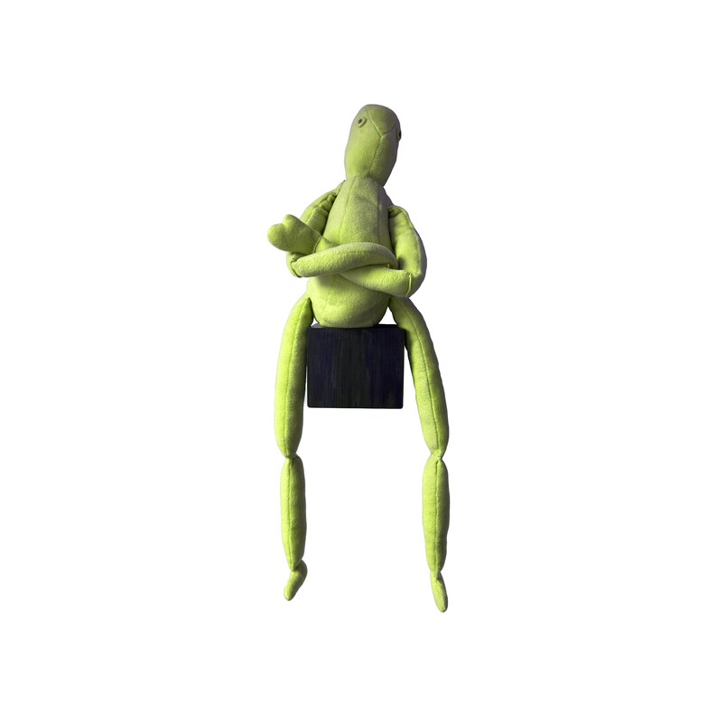 May include: A lime green fabric doll with a seated pose. The doll's arms are crossed, and it sits atop a small, dark wooden block. The doll's legs dangle, and the background is white.