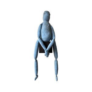 May include: A light blue fabric doll with a minimalist design. The doll is seated on a small, dark brown wooden block. Its arms and legs are long and slender, and it has a simple, rounded head.
