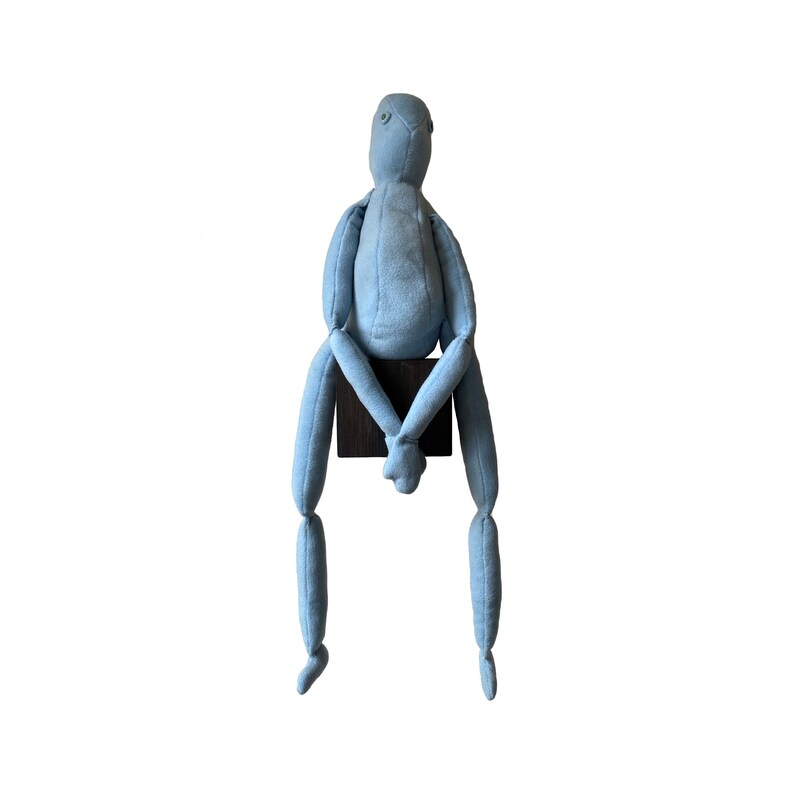 May include: A light blue fabric doll with a minimalist design. The doll is seated on a small, dark brown wooden block. Its arms and legs are long and slender, and it has a simple, rounded head.