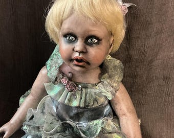 May include: A doll with blonde hair, pale skin, and dark eye makeup, wearing a ruffled, iridescent blue and green top with small pink rose accents and matching blue bloomers. The doll has a distressed appearance with smudged facial features.