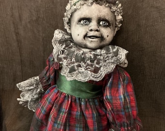 May include: A doll with a pale, weathered face, bright blue eyes, and dark shading around the eyes and mouth. It wears a red, green, and blue plaid dress with a green satin waistband, a white lace collar, lace-trimmed sleeves, and a matching plaid bow.