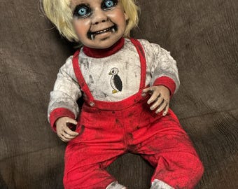 May include: A doll with blonde hair, bright blue eyes, and visible fangs. It wears a long-sleeved white shirt featuring a small penguin graphic and red overalls with suspenders. The doll's face, hands, and feet have a distressed, dirty appearance.
