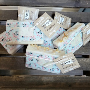 May include: A stack of four handmade soap bars wrapped in floral fabric with brown paper tags that read "All Natural Soap 1907 Calendula Broken Arrow, Oklahoma".