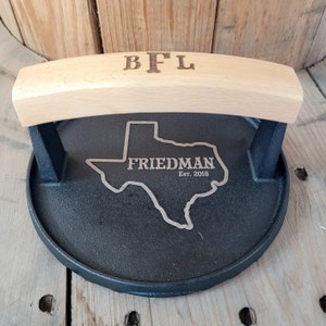 May include: A black cast iron grill press with a light wood handle. The handle is engraved with the initials "BFL". The press features an engraved outline of the state of Texas with the name "FRIEDMAN" and the year "Est. 2018".