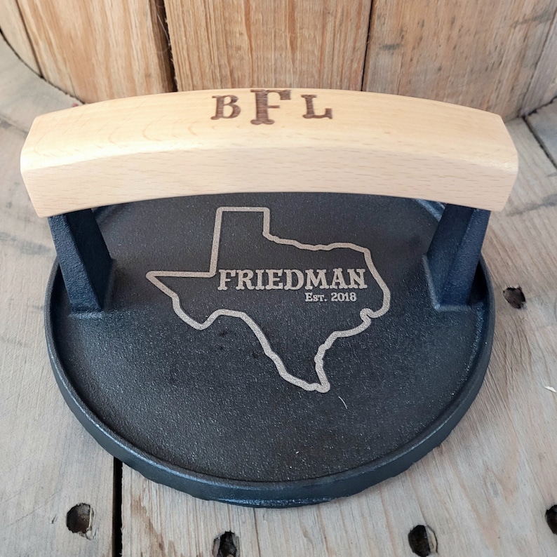 May include: A black cast iron grill press with a light wood handle. The handle is engraved with the initials "BFL". The press features an engraved outline of the state of Texas with the name "FRIEDMAN" and the year "Est. 2018".