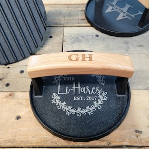 May include: A black cast iron grill press with a light wood handle engraved with "GH". The press has the text "The Li-Hares EST. 2017" in a decorative wreath design. The press is on a wooden surface.