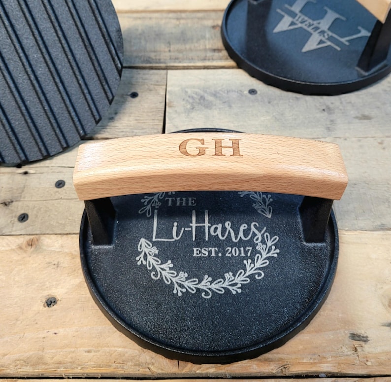 May include: A black cast iron grill press with a light wood handle engraved with "GH". The press has the text "The Li-Hares EST. 2017" in a decorative wreath design. The press is on a wooden surface.
