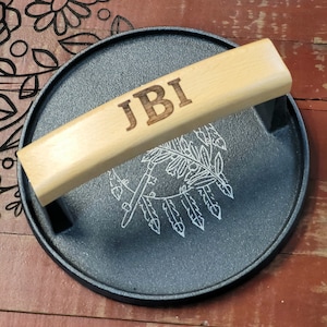 May include: A round, black cast iron serving tray with a light wood handle. The handle is engraved with the initials "JBI". The tray has a white etched design of feathers and arrows.
