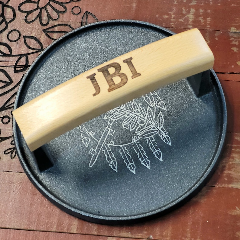 May include: A round, black cast iron serving tray with a light wood handle. The handle is engraved with the initials "JBI". The tray has a white etched design of feathers and arrows.