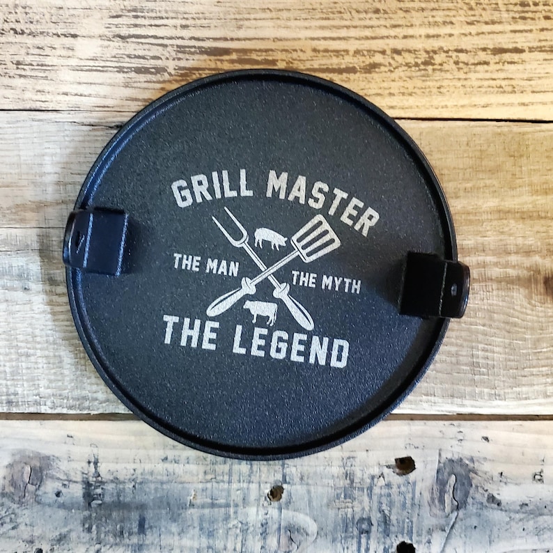 May include: A circular, dark blue cast iron grill lid with the text "GRILL MASTER THE MAN THE MYTH THE LEGEND" in white. The lid has crossed grilling tools and images of a pig and cow. Two black handles are attached.