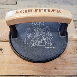 May include: A black cast iron burger press with a light wood handle engraved with the name "SCHLITTLER". The press features a detailed engraved crest and the date "9-15-2018".