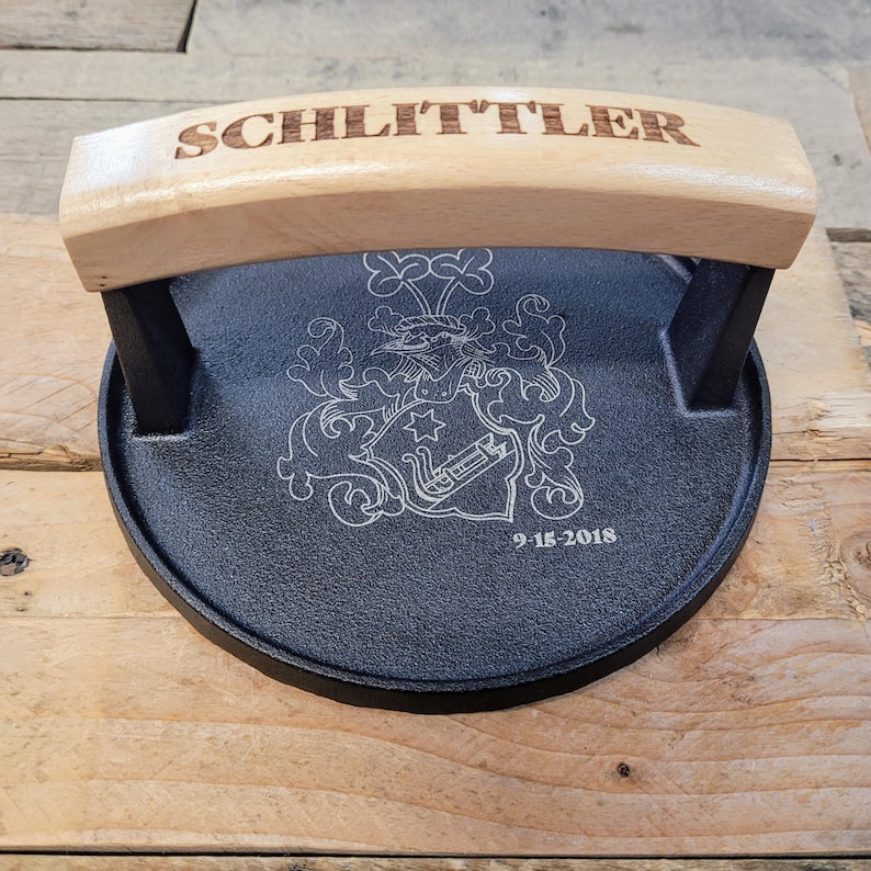May include: A black cast iron burger press with a light wood handle engraved with the name "SCHLITTLER". The press features a detailed engraved crest and the date "9-15-2018".