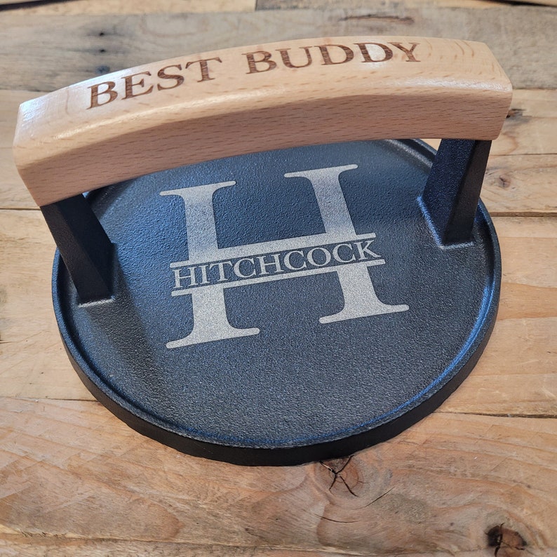 May include: A black cast iron burger press with a light wood handle. The handle is engraved with "BEST BUDDY". The press features the name "HITCHCOCK" with a large letter "H" in the center. The press is designed for making burgers.