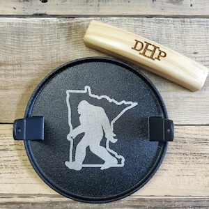 May include: A black metal ashtray featuring a Bigfoot silhouette within the outline of Minnesota. A wooden piece with the initials "DHP" is next to the ashtray. The ashtray has two black metal holders.