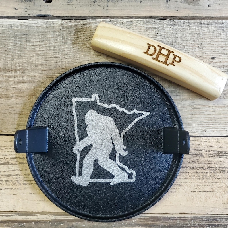May include: A black metal ashtray featuring a Bigfoot silhouette within the outline of Minnesota. A wooden piece with the initials "DHP" is next to the ashtray. The ashtray has two black metal holders.