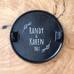 May include: A black, circular ashtray featuring the names "Randy & Karen" in white script. The ashtray has two built-in rests and is decorated with sprigs of leaves. The Roman numeral "NII" is below the names.
