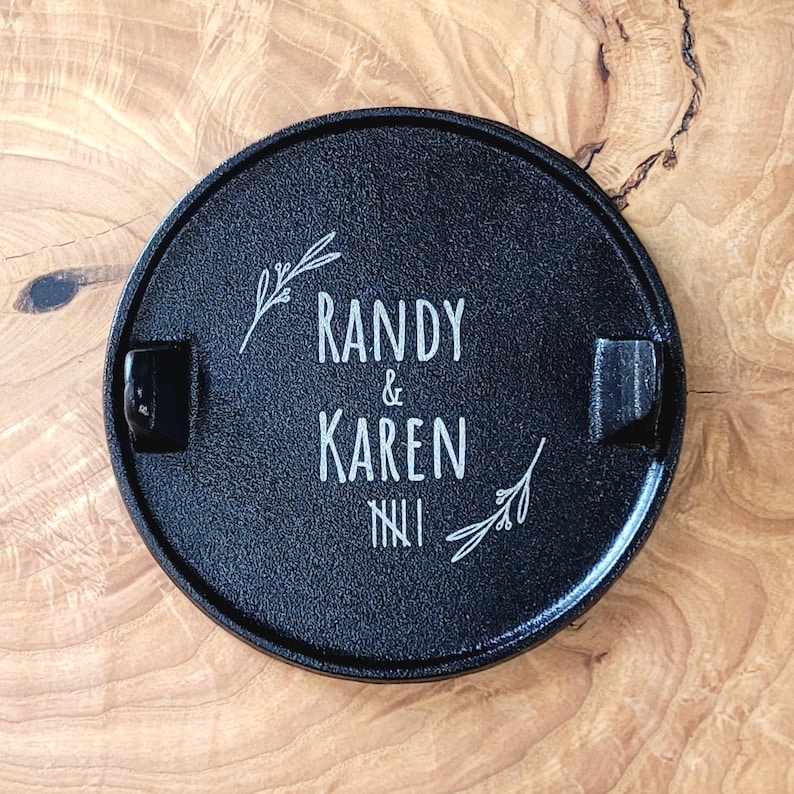 May include: A black, circular ashtray featuring the names "Randy & Karen" in white script. The ashtray has two built-in rests and is decorated with sprigs of leaves. The Roman numeral "NII" is below the names.