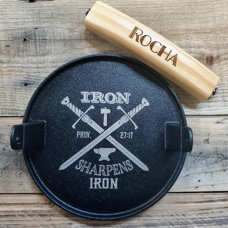 May include: A black cast iron lid with the text "IRON SHARPENS IRON" and crossed swords. A wooden handle with the name "ROCHA" engraved on it rests on the lid. The lid has two handles and is on a wooden surface.