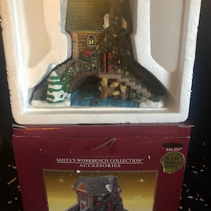 May include: A porcelain Santa's Workshop Collection accessory, a small cottage with a bridge and a stream, in a red box with the text "Santa's Workbench Collection Accessories" and "New 2000 Design".
