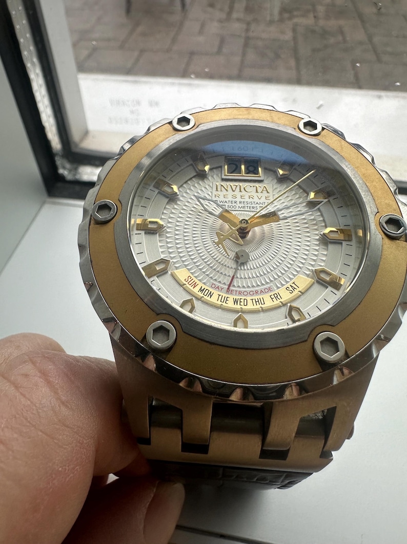 May include: A gold and silver Invicta Reserve watch with a white face and a day retrograde feature. The watch is water resistant to 500 meters.