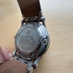 May include: A silver Invicta Reserve watch with a brown leather band. The watch face is not visible, but the back of the watch is visible, showing the brand name and model number. The watch is on a wooden surface.