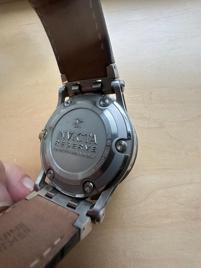 May include: A silver Invicta Reserve watch with a brown leather band. The watch face is not visible, but the back of the watch is visible, showing the brand name and model number. The watch is on a wooden surface.