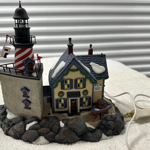 May include: A decorative lighthouse figurine with a red and white striped tower, a black top, and a small house. The base is made to look like rocks with snow. A small boat is next to the house. The figurine has a power cord.