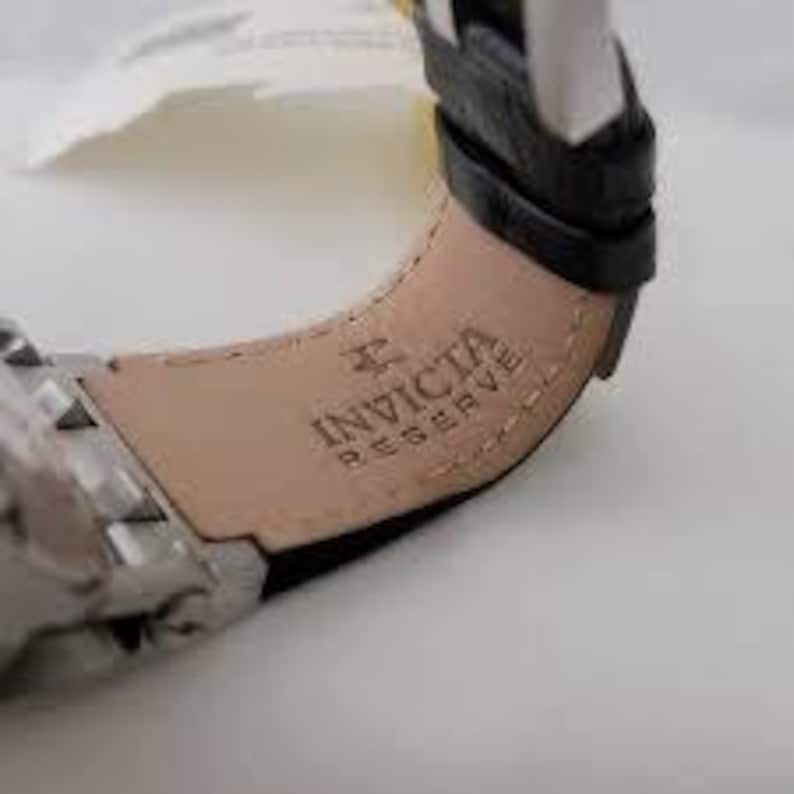 May include: Close-up of a black leather watch band with the Invicta Reserve logo embossed in silver.