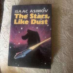 Könnte beinhalten: Ein Buchcover mit einem lila und schwarzen Hintergrund, auf dem ein Raumschiff durch den Weltraum fliegt. Der Titel des Buches lautet "The Stars, Like Dust" von Isaac Asimov.