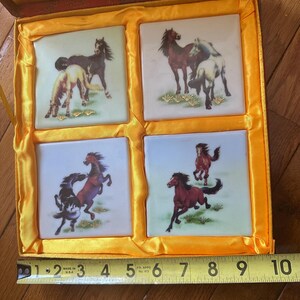 May include: A wooden box with a yellow satin lining contains four square ceramic tiles. Each tile features a different image of horses in various poses, painted in shades of brown, black, and white against a white background. The tiles are surrounded by a yellow border.