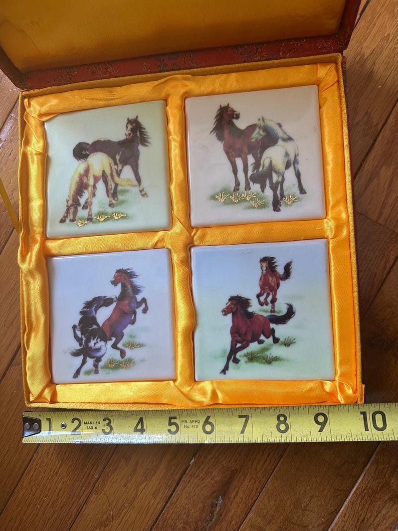 May include: A wooden box with a yellow satin lining contains four square ceramic tiles. Each tile features a different image of horses in various poses, painted in shades of brown, black, and white against a white background. The tiles are surrounded by a yellow border.