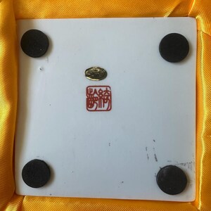 May include: A white ceramic tile with four black rubber feet on the bottom. The tile has a gold seal with Chinese characters and a red stamp with Chinese characters. The tile is in a yellow satin box.