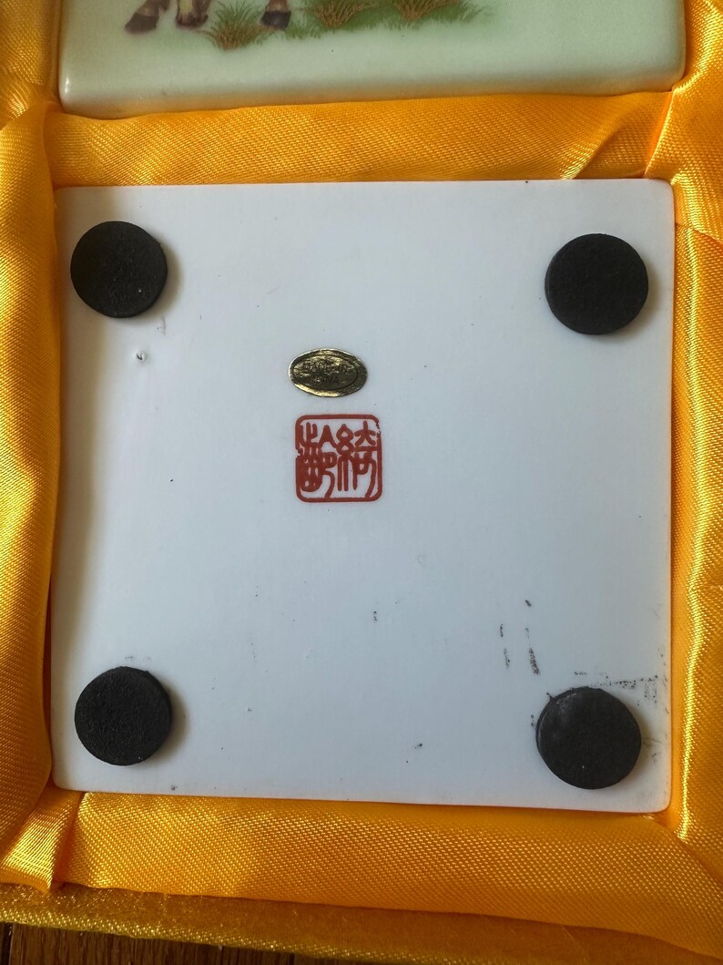 May include: A white ceramic tile with four black rubber feet on the bottom. The tile has a gold seal with Chinese characters and a red stamp with Chinese characters. The tile is in a yellow satin box.