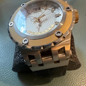 May include: A gold and silver Invicta watch with a white face and a day retrograde feature. The watch has a black leather band.