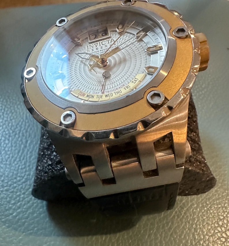 May include: A gold and silver Invicta watch with a white face and a day retrograde feature. The watch has a black leather band.