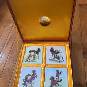 May include: A red and gold box with a yellow interior. The box contains four square tiles with images of horses on them. The tiles are white with a blue background and gold accents.