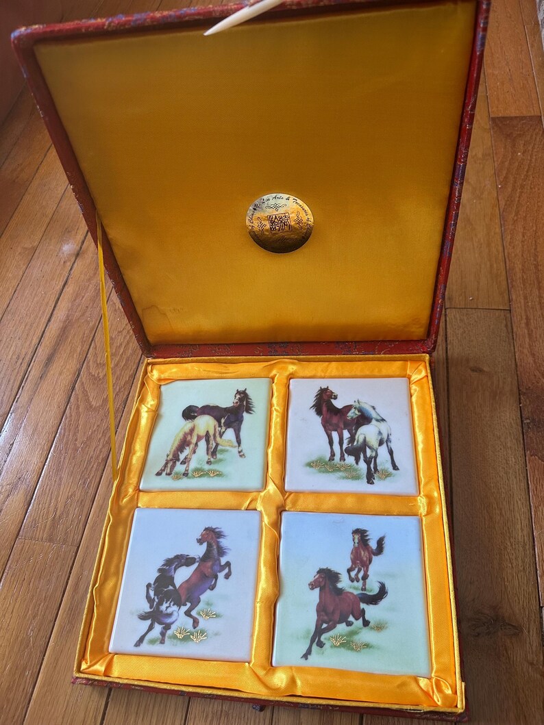 May include: A red and gold box with a yellow interior. The box contains four square tiles with images of horses on them. The tiles are white with a blue background and gold accents.