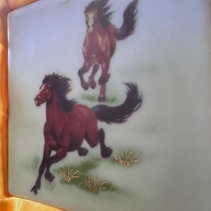May include: Two brown horses running on a green field with gold accents. The horses are painted on a light blue ceramic tile.
