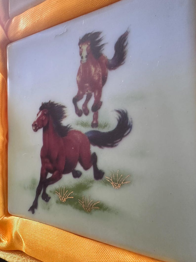 May include: Two brown horses running on a green field with gold accents. The horses are painted on a light blue ceramic tile.