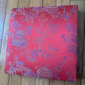 May include: A red box with a gold and purple floral pattern. The box has a gold trim and a gold tassel.