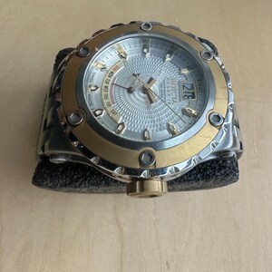 May include: A silver and gold Invicta watch with a white face and multiple subdials. The watch has a black rubber band and a date window at the 3 o'clock position.