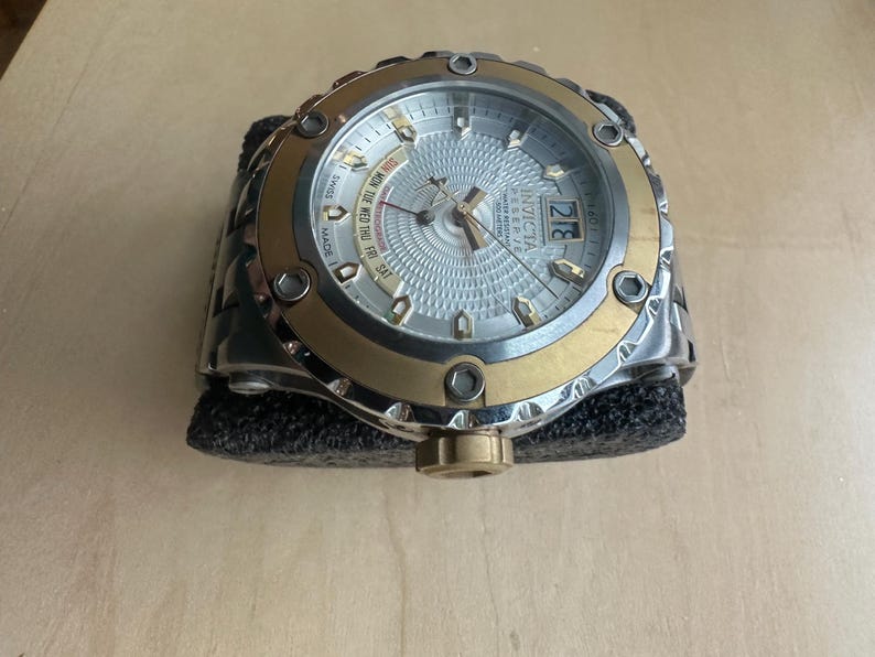 May include: A silver and gold Invicta watch with a white face and multiple subdials. The watch has a black rubber band and a date window at the 3 o'clock position.