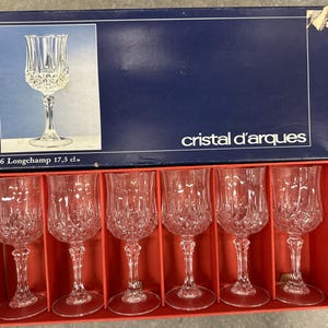 May include: A set of six clear glass stemware pieces in a blue and red cardboard box labeled Cristal d'Arques 6 Longchamp 17.5 cl. Each glass features a faceted diamond-cut pattern on the lower bowl, a slender stem, and a circular base.